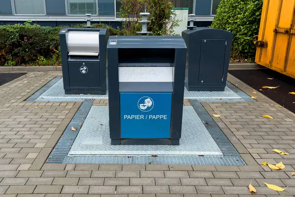 Kağıt atığı için tasarlanmış modern bir şehir parkında üç geri dönüşüm kutusu bulunuyor. Bölge temiz, yeşilliklerle çevrili ve taşlarla döşenmiş..