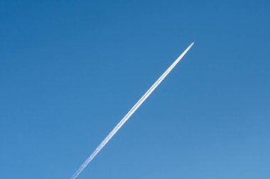 Bir jet parlak mavi gökyüzüne karşı uzun beyaz bir iz bırakır, çarpıcı bir kontrast yaratır. Gün ışığı atmosferin berraklığını ve derinliğini vurgular..