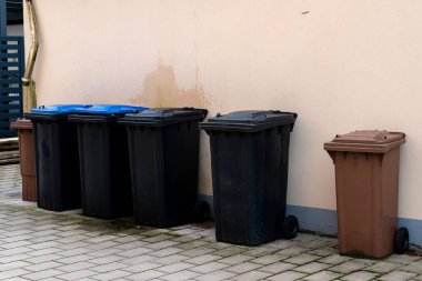 Farklı renklerde çöp bidonları bina duvarına karşı dizilmiş. Bölge iyi korunmuş görünüyor, temiz ve düzenli bir ortam var..