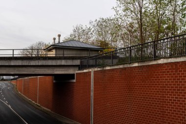 İki şeritli boş bir yola paralel giden tuğladan bir duvar, üzerinde köprü var. Yeşillik bir sanayi binasını çevreleyerek öğleden sonra sakin bir şehir ortamı yaratır..