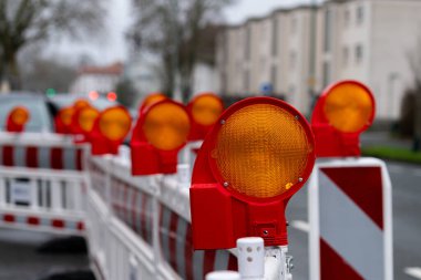Kalabalık bir caddede inşaat bariyerlerinin üzerindeki parlak turuncu uyarı ışıkları sürücüleri alarma geçiriyor. Sahne, şehir altyapı çalışmalarının altını çizerek akşam saatlerinde bir anı yakalıyor..