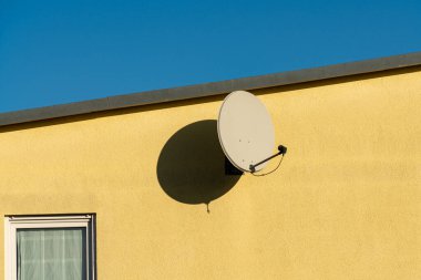 Açık mavi bir gökyüzünün altında parlak sarı bir duvara büyük bir uydu anteni monte edilir. Çanak duvara gölge düşürüyor, görsel ilgi ve kontrastlık yaratıyor..