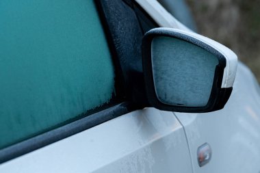 Donmuş aynalı beyaz bir araba serin bir çevrede park edilmiş duruyor. Buzlu koşullar, cam bir buz tabakası gösterirken ve etrafındaki bölge soğuk görünürken belirgindir..