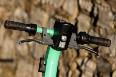 Yeşil e-scooter gidonlarının yakın görüntüsü, kabzaları ve kontrol kollarını vurguluyor. Arka planda doğal taş dokuları var, canlı renkle çarpıcı bir kontrast oluşturuyor..