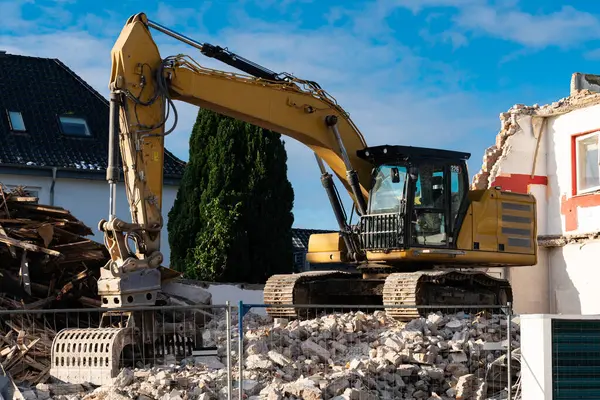 Hasarlı bir binanın kalıntılarını parçalayan bir kazıcı inşaat alanında çalışıyor. Güneş ışığı bölgeyi aydınlatıyor, molozların arasındaki makineleri vurguluyor..