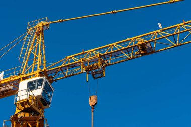 Büyük sarı bir vinç açık mavi bir gökyüzüne karşı dimdik ayakta duruyor. Operatörün kulübesi görülebiliyor, aşağı sarkan bir kancayla birlikte, bir inşaat alanında ağır kaldırmaya hazır..