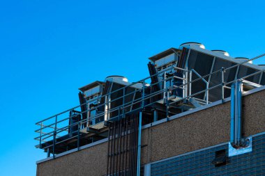 Modern bir binanın çatısına büyük klima birimleri kurulur. Parlak mavi bir gökyüzünün arka planında oturuyorlar, kentsel mimaride endüstriyel tasarımı bir anlığına sunuyorlar..