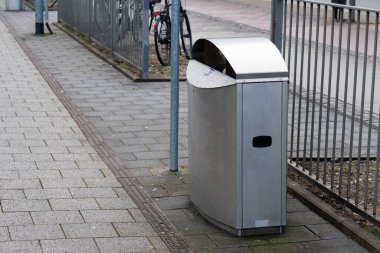 Metal bir çitin yanındaki kaldırımda duran gümüş bir çöp tenekesi. Bisikletler yakınlarda park edilmiş. Sahne temiz kaldırımlı bir kentsel alan, düşünceli şehir planlamasını gösteriyor..