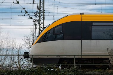 Parlak sarı önlü parlak bir tren raylar boyunca sorunsuzca ilerliyor. Arka planda, uzun elektrik direkleri bulutlu bir gökyüzüne doğru uzanıyor ve ilgi çekici bir şehir manzarası yaratıyor..