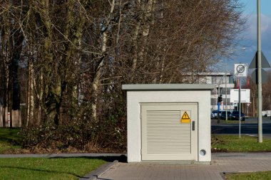 Bir sandık, kentsel alanda ağaçlarla çevrili bir yolun kenarında huzur içinde duruyor. Açık mavi gökyüzü arkaplanına karşı sağlam inşaat ve güvenlik uyarı sembolünü sergiliyor..