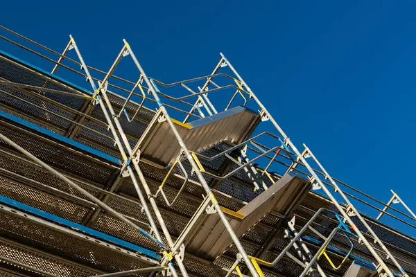 Çoklu katmanlı modern iskele canlı mavi bir gökyüzüne karşı konumlandırılmıştır. Yapıda metal korkuluklar ve merdivenler yer alıyor, inşaat faaliyetleri vurgulanıyor.