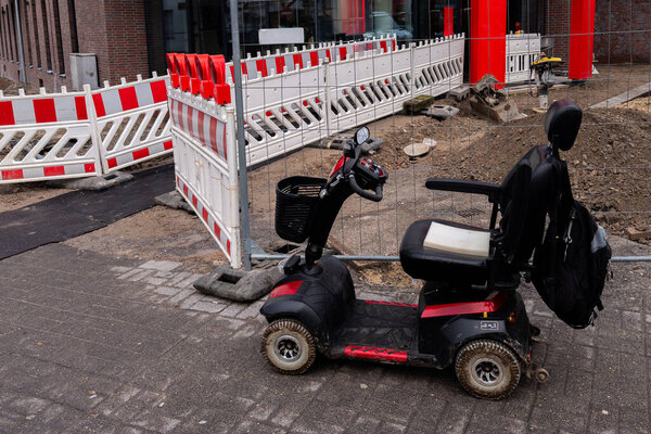 A mobility scooter parked beside a construction zone features barriers and an unfinished surface. The setting reflects ongoing urban development with some materials scattered around.