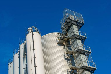 Büyük endüstriyel depolama siloları parlak mavi bir gökyüzüne karşı dik dururlar. Metal yapılarda merdiven ve korkuluklar var. Bu da aşağıda güneş ışığıyla dolu bir imalat ortamı olduğunu gösteriyor..