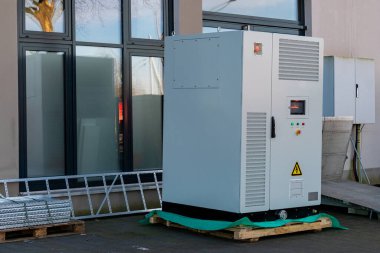 Çağdaş bir binanın dışında büyük bir endüstriyel soğutma ünitesi yeşil bir minderin üzerinde duruyor. Birim kontrol panelleri, uyarı işaretleri ve metal yapı ve pencerelerle çevrilidir..