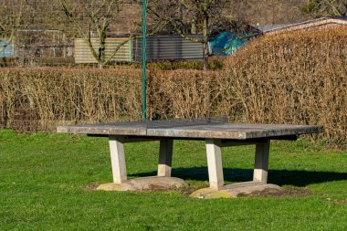 Güçlü bir beton piknik masası yemyeşil bir parkta duruyor, yemyeşil çimenler ve çitlerle çevrili. Güneş ışığı masa yüzeyine vurgu yaparak ziyaretçileri açık hava yemekleri ve toplantıların tadını çıkarmaya davet ediyor..