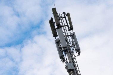 Yüksek bir baz istasyonu, beyaz bulutlarla dolu bir gökyüzüne uzanan antenlerle belirgin bir şekilde yükselir. Yapı, şehir ortamında modern iletişim teknolojisini simgeliyor..