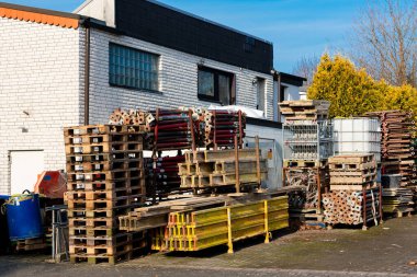 Depoda bir yığın ahşap palet ve inşaat iskelesi bulunuyor. Parlak güneş ışığı düzenli düzenlemeyi vurguluyor. Arkaplanda bir bina ve yeşillik yer alıyor.