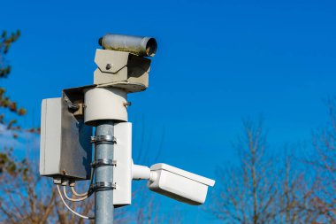 Bölgeyi gözetleyen bir güvenlik kamerası bir direğe yerleştirilmiş. Cihaz aşağıdaki faaliyetleri izlemeyi amaçlıyor, açık mavi gökyüzü arkaplanı sağlıyor.