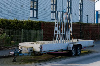 Bir binanın yakınında bir araba römorku duruyor. Römork basit bir çerçeve ile donatılmıştır, gün ışığında çit boyunca yeşil alan bir caddede yer almaktadır..