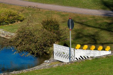 Bir su kontrol yapısı, yansıtıcı bir yüzeye sahip sakin bir gölet tarafından görülebilir. Çevredeki yemyeşil çimenlerin ve ağaçların arka planında sarı güvenlik bariyerleri belirgin şekilde duruyor.