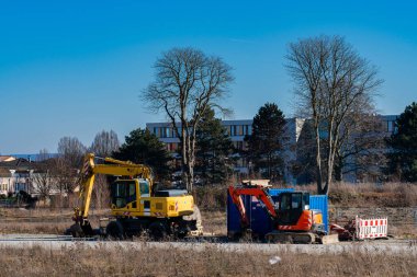 Kazı makineleri bir inşaat sahasında modern bir binanın yakınındaki toprağı kazıp taşımakla meşguller. Bölge açık ve açık bir ağaç manzarası ve mavi gökyüzü var..