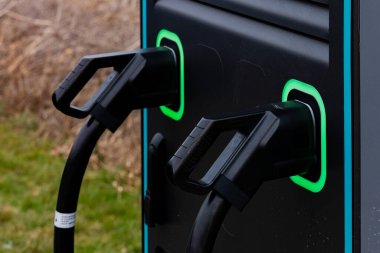 Çimenli bir bölgedeki bir elektrikli araç şarj istasyonunda iki şarj konnektörü hazır bekliyor. Canlı yeşil ışıklar istasyonun kullanıma hazır ve kullanılabilir olduğunu gösteriyor..