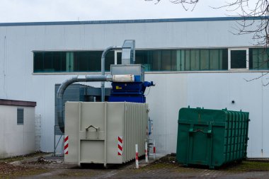 Fabrika binasının dışında egzoz boruları bağlı büyük bir endüstriyel makine kuruluyor. Yakınlarda yeşil bir atık konteyneri bulunur ve olay yeri kış atmosferiyle karakterize edilir..