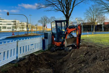 Küçük bir kazıcı, yol kenarındaki bir alanda inşaat için hendek kazıyor. Ağaçların ve binaların etrafında açık gökyüzü ile güneşli bir günü yansıtan kentsel bir zemin oluşturur..