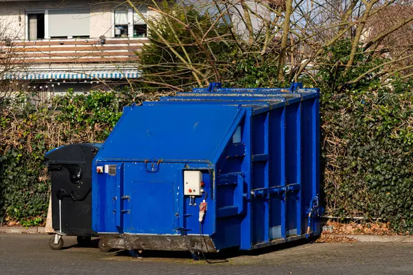 Bir yerleşim bölgesinde siyah bir çöp kutusunun yanında mavi bir çöp konteynırı duruyor. Çevreleyen çalılar ve ağaçlar doğal yeşillik sağlar, kentsel çevre ile çelişir..