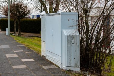 Sakin bir banliyö mahallesinde bir yürüyüş yolunun yanına bir çift elektrik hizmet kutusu yerleştirildi. Sahnede çıplak ağaçlar ve iyi bakımlı çimenler yer alıyor ve bu da baharın başlarında.