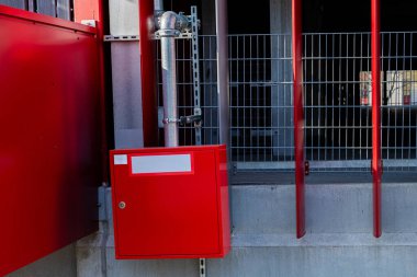 Kırmızı bir yangın alarmı bir kapının yanındaki beton duvara güvenli bir şekilde monte edilir. Kutunun arkasında metal parmaklıklar çitle çevrili bir alan oluşturarak acil durum müdahaleleri için güvenli ve düzenli bir ortam oluşturuyor..