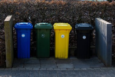 Mavi, yeşil, sarı ve siyah renkli dört farklı çöp bidonu, bir çitle çevrili bir taş yolun yanında düzgün bir şekilde sıralanmış, bu da topluluğun geri dönüşüm ve temizliğe olan bağlılığını gösteriyor..