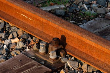 Paslı metal ve çakıllı yıpranmış bir demiryolu hattına yakın çekim. Güneş ışığı, öğleden sonra geç saatlerde yüzeylere sıcak tonlar dökerek dokuları ve yaşlanma materyallerini vurgular..