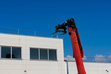 Kırmızı bir forklift kolunu modern bir binanın yapım aşamasındaki üst kısmına doğru uzatır. İşçiler bir sonraki aşamaya hazırlanırken açık mavi gökyüzü çarpıcı bir zemin sağlıyor..