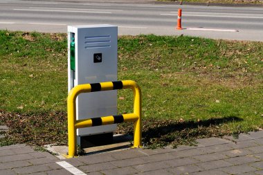 Modern bir elektrikli taşıt park yeri tasarlaması işlek bir yolun yakınındaki iyi bakımlı bir çimenliğin yanında duruyor. Sarı bir kazık ödeme ekipmanlarına koruma sağlar ve güvenliği arttırır..