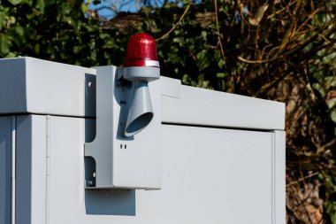 Açık havada duran bir elektrik kabininde parlak bir kırmızı uyarı ışığı ve yanında da bir boynuz var. Arkaplan açık mavi gökyüzünün altında yeşil sarmaşıklarla dolu..