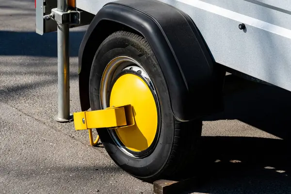 Parlak sarı bir tekerlek kıskacı park halindeki bir aracın lastiğine sabitlendi. Ayarda, aracın gün boyunca hareketsiz kaldığını gösteren, berrak görüş açısına sahip asfalt bir cadde yer alıyor..