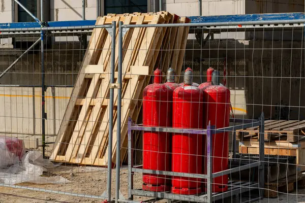 Tahta paletler duvara yaslanırken, kırmızı gaz tüpleri inşaat alanındaki metal bir çitin arkasında güvenceye alınır. Bu organize sanayi bölgesinde güvenlik tedbirleri belirgin.