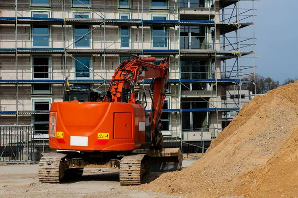 Turuncu bir kazıcı inşaat sahasında faaliyet gösteriyor, kısmen inşa edilmiş bir yapı yakınındaki toprak ve enkazı hareket ettiriyor. Arkaplanda işçiler mevcut.