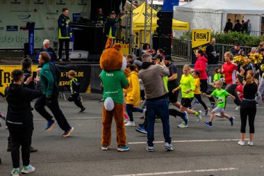 ALMANY, PADERBORN, 77 EASTER RUN, 04-19-2025. Çeşitli yaşlardan koşucular, büyük bir kalabalığın alkışladığı canlı bir yarışa katılıyorlar. Kahverengi kostümlü bir maskot heyecan katar. Sadece editör kullanımı.
