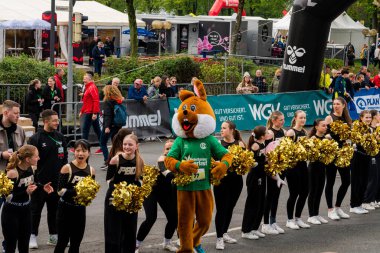 ALMANY, PADERBORN, 77 EASTER RUN, 04-19-2025. Yeşil elbiseli neşeli bir maskot canlı bir spor etkinliğinde ponpon kızların yanında yürüyor. Sadece editör kullanımı.