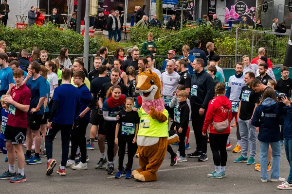 ALMANY, PADERBORN, 77 EASTER RUN, 04-19-2025. Çok sayıda katılımcı kentsel bir ortamda koşuya hazırlanıyor. Kostümlü neşeli bir maskot, koşucularla etkileşime girer. Sadece editör kullanımı.
