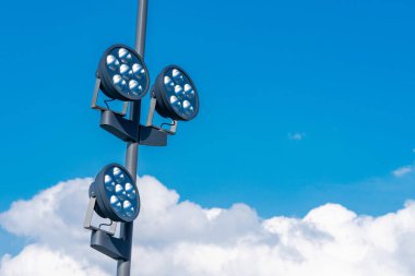 Projektörler uzun bir direğe kurulur. Mavi, parlak bir gökyüzü ve birkaç tüylü bulut. Işıklar üç kümeye ayrılmıştır, açık hava aktiviteleri için aydınlatma sağlanmaktadır..