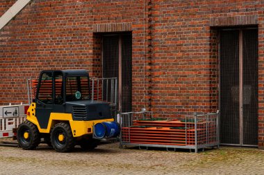Tuğla bir binanın yanına küçük bir forklift park edilmiş. Aracın yanında yığılmış metal borular ve inşaat malzemeleri var. Bu da bölgede çalışmaların devam ettiğini gösteriyor..