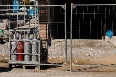 Bir inşaat sahasında tel örgülerin arkasında güvenli bir şekilde depolanmış birkaç gaz silindiri bulunur. Bölgede bitmemiş yapılar ve inşaat malzemeleri gösteriliyor, devam eden çalışmalar vurgulandı.