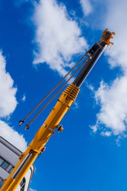 Uzun bir vinç kolu yukarı uzanır ve mavi gökyüzüne karşı sarı rengini gösterir. Sahne inşaat araçlarını yakalıyor, kentsel bir yerde devam eden çalışmaları vurguluyor.