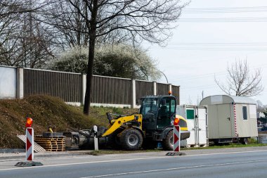 İnşaat ekipmanları işlek bir yolun yanında yer alıyor. Sarı makineler ahşap paletlerin yanında duruyor ve portatif bir ofis, açık gökyüzü ve arka planda seyrek ağaçlar..