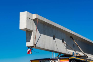 Gün ışığında büyük bir beton kiriş düz bir kamyona kaldırılıyor. Sahne, altyapı geliştirmede kullanılan inşaat malzemelerinin ölçeğini vurguluyor. Açık mavi gökyüzü tepemizde.