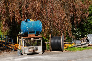 Makineler ve malzemeler gün ışığında açık bir alanda toplanıyor. Büyük bir kablo makarasının yanında mavi bir su tankı ve inşaat malzemeleri bulunuyor..