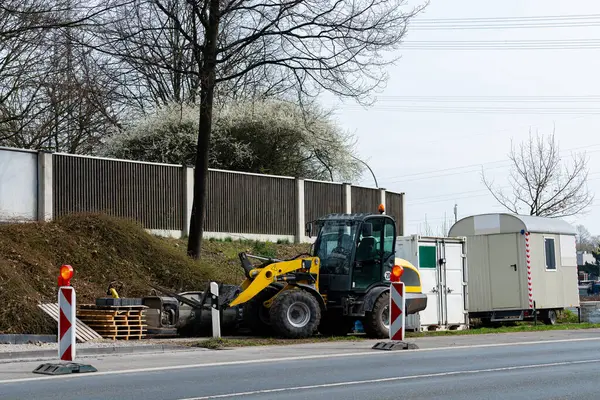 İnşaat ekipmanları işlek bir yolun yanında yer alıyor. Sarı makineler ahşap paletlerin yanında duruyor ve portatif bir ofis, açık gökyüzü ve arka planda seyrek ağaçlar..